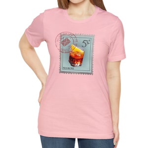 May include: A pink t-shirt with a vintage-style stamp featuring a glass of Negroni with an orange slice. The stamp reads "5c" and "NEGRONI".