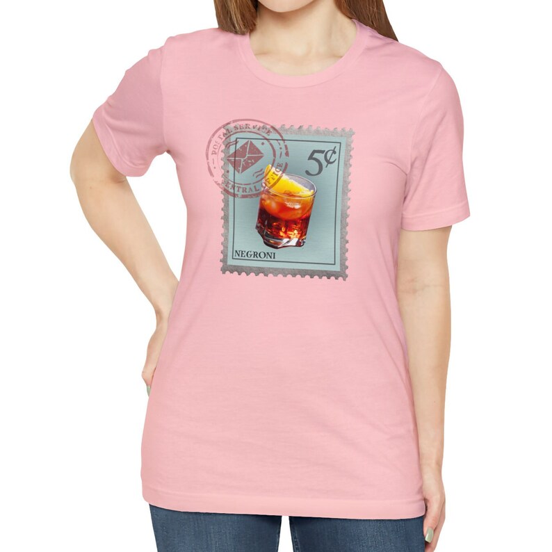 May include: A pink t-shirt with a vintage-style stamp featuring a glass of Negroni with an orange slice. The stamp reads "5c" and "NEGRONI".