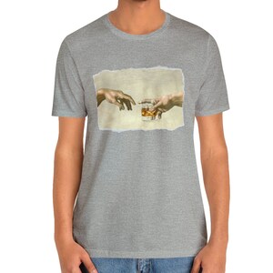 K&ouml;nnte beinhalten: Ein graues T-Shirt mit einem Aufdruck von zwei H&auml;nden, die sich gegenseitig erreichen und ein Glas Whisky halten. Der Druck ist auf einem strukturierten, gebrochenen Wei&szlig;-Hintergrund.