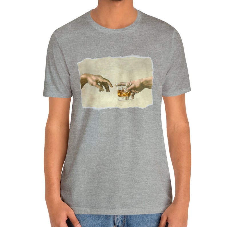 K&ouml;nnte beinhalten: Ein graues T-Shirt mit einem Aufdruck von zwei H&auml;nden, die sich gegenseitig erreichen und ein Glas Whisky halten. Der Druck ist auf einem strukturierten, gebrochenen Wei&szlig;-Hintergrund.