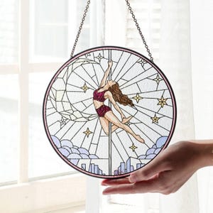 Pode incluir: Obra de arte em vitral circular com uma mulher a dançar numa barra. O design inclui uma paisagem urbana, lua, estrelas e nuvens. A mulher usa um traje bordô. Borda rosa e suspensão por corrente.