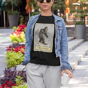 Pode incluir: Uma camiseta preta com uma ilustra&ccedil;&atilde;o de estilo vintage do P&eacute; Grande em um cart&atilde;o com o texto "The Bigfoot".