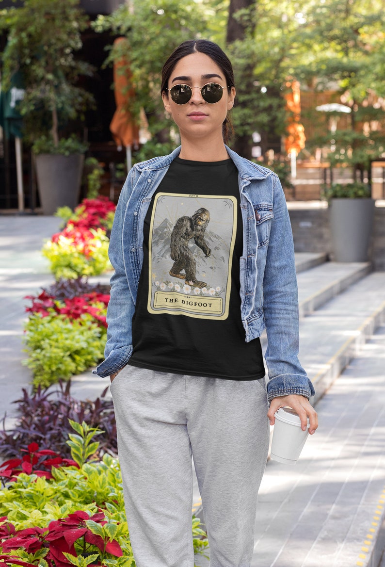 Pode incluir: Uma camiseta preta com uma ilustra&ccedil;&atilde;o de estilo vintage do P&eacute; Grande em um cart&atilde;o com o texto "The Bigfoot".