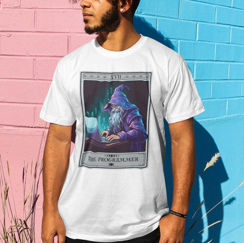 May include: White t-shirt with a graphic of a wizard with a long white beard, wearing a purple robe and a pointed hat, sitting at a desk and typing on a laptop. The graphic is in a card-like frame with the text "XVII THE PROGRAMMER" at the bottom.