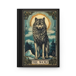 May include: A black journal featuring a detailed illustration of a wolf in a forest setting, with a sun and clouds in the background. The words "THE WOLF" are at the bottom of the image. The artwork has a vintage, art nouveau style.