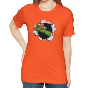 May include: Orange t-shirt with a graphic of a green zombie hand reaching out of a ripped hole in the fabric, holding a brown cup with a straw.