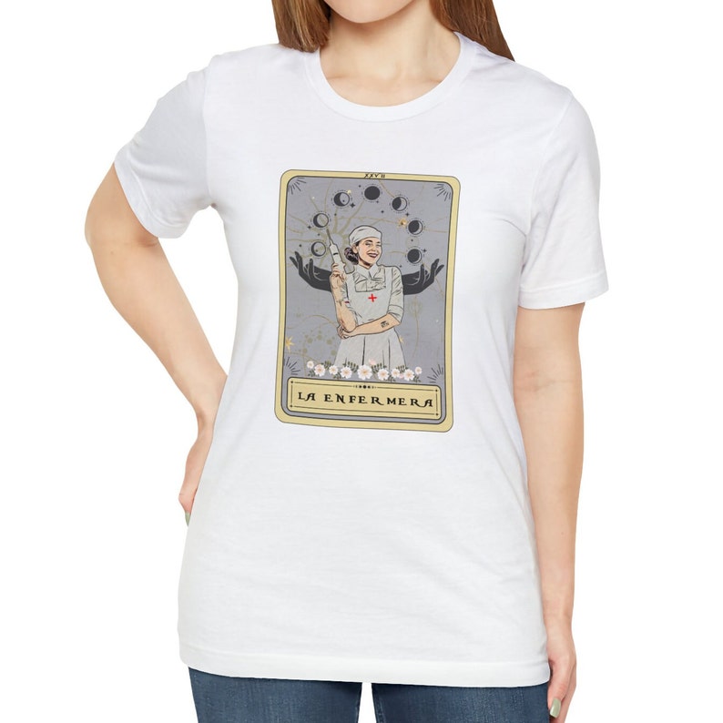 May include: White t-shirt featuring a tarot card design with a woman in a nurse's uniform holding a syringe. The card is titled "La Enfermera" in a gold frame.