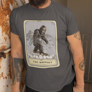 Pode incluir: Uma camiseta cinza escura com uma ilustra&ccedil;&atilde;o vintage de um P&eacute; Grande em um cart&atilde;o. O cart&atilde;o tem o texto "XXV II" e "THE BIGFOOT".