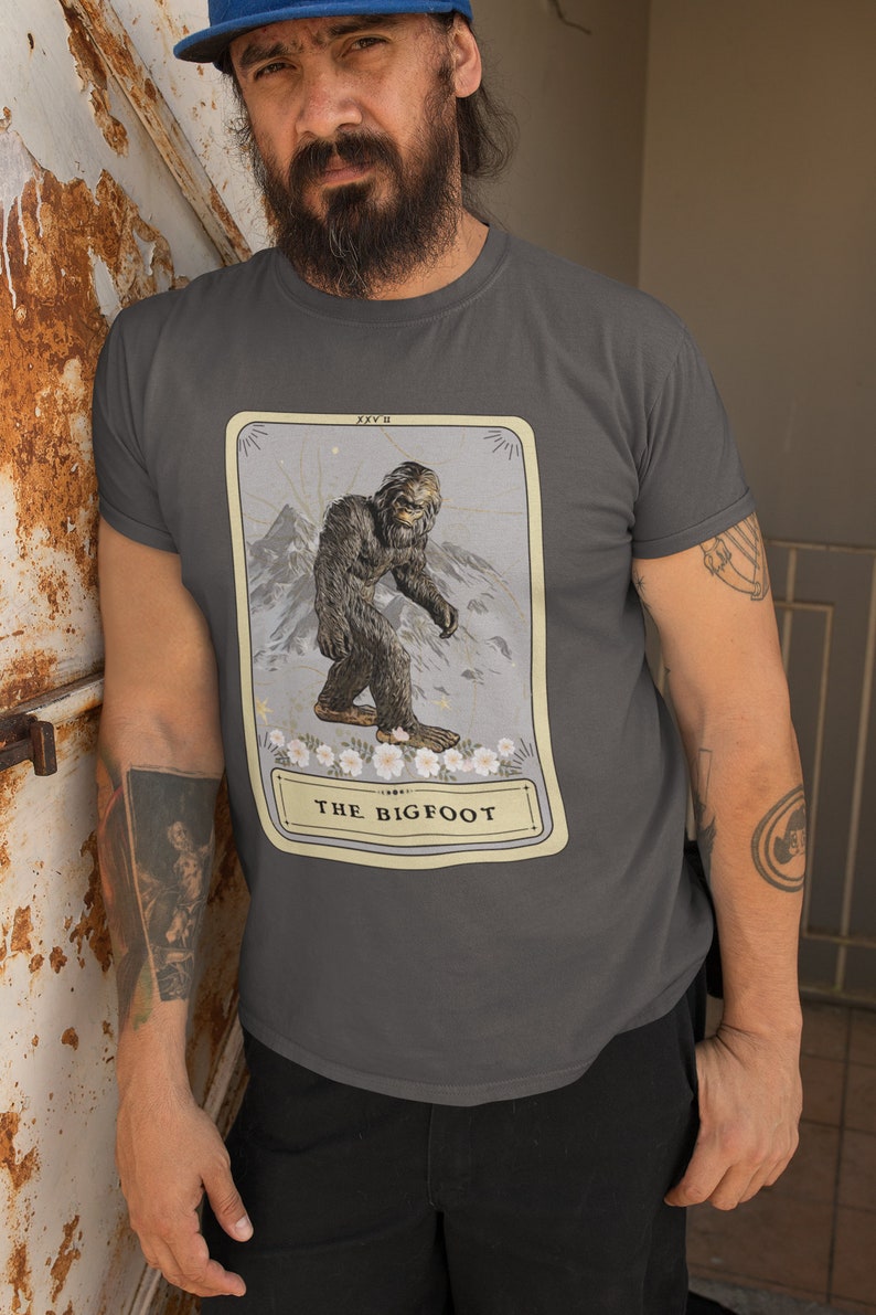 Pode incluir: Uma camiseta cinza escura com uma ilustra&ccedil;&atilde;o vintage de um P&eacute; Grande em um cart&atilde;o. O cart&atilde;o tem o texto "XXV II" e "THE BIGFOOT".