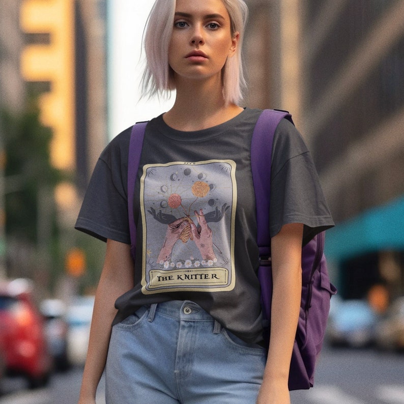 May include: A dark gray t-shirt with a graphic of a tarot card featuring a pair of hands knitting with a moon and stars design. The text on the card reads "The Knitter".