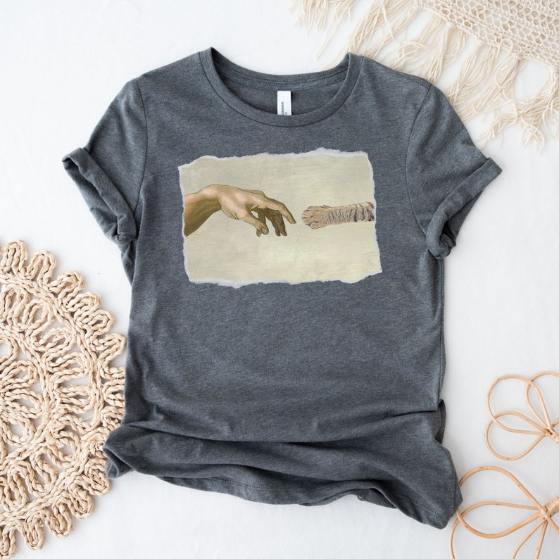 May include: A dark grey t-shirt with a print of a cat paw and a human hand reaching towards each other, inspired by the famous "The Creation of Adam" painting.