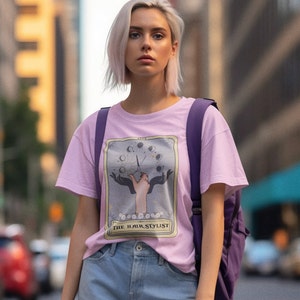 Puede incluir: Una camiseta rosa con un gr&aacute;fico de una mano sosteniendo tijeras frente a un dise&ntilde;o de fases lunares. El texto "THE HAIR STYLIST" est&aacute; impreso debajo del gr&aacute;fico.