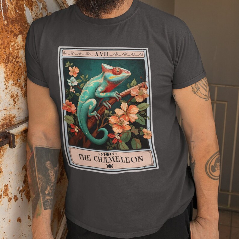 May include: A dark gray t-shirt with a graphic of a tarot card featuring a green and blue chameleon on a branch with pink flowers. The card is labeled "XVIII THE CHAMELEON".