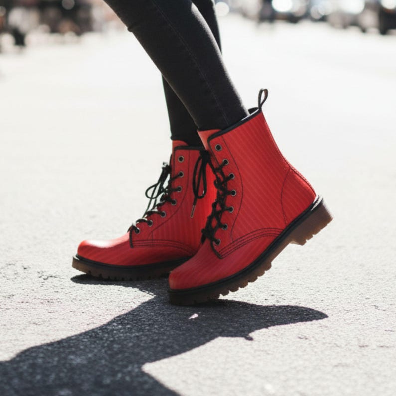May include: Bright red combat boots with black laces and a textured pattern. The boots have a brown sole and are worn with black pants. The image is taken outdoors on a sunny day.
