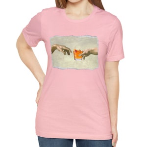 May include: A pink t-shirt with a graphic of the "Creation of Adam" painting, but instead of God touching Adam's finger, they are both reaching for a glass of orange juice with a cherry.