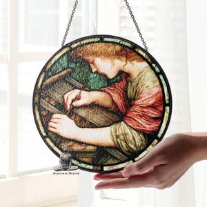 Peut inclure: Vitrail rond représentant une femme tissant, avec un cadre en métal noir et une chaîne de suspension. L'œuvre d'art utilise des tons rouges, verts et bruns. L'image est tenue dans une main.