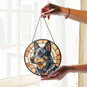 May include: Round stained glass panel featuring a blue heeler dog portrait. The dog's head is centered, surrounded by a geometric pattern in shades of orange and brown. A metal chain is attached for hanging.