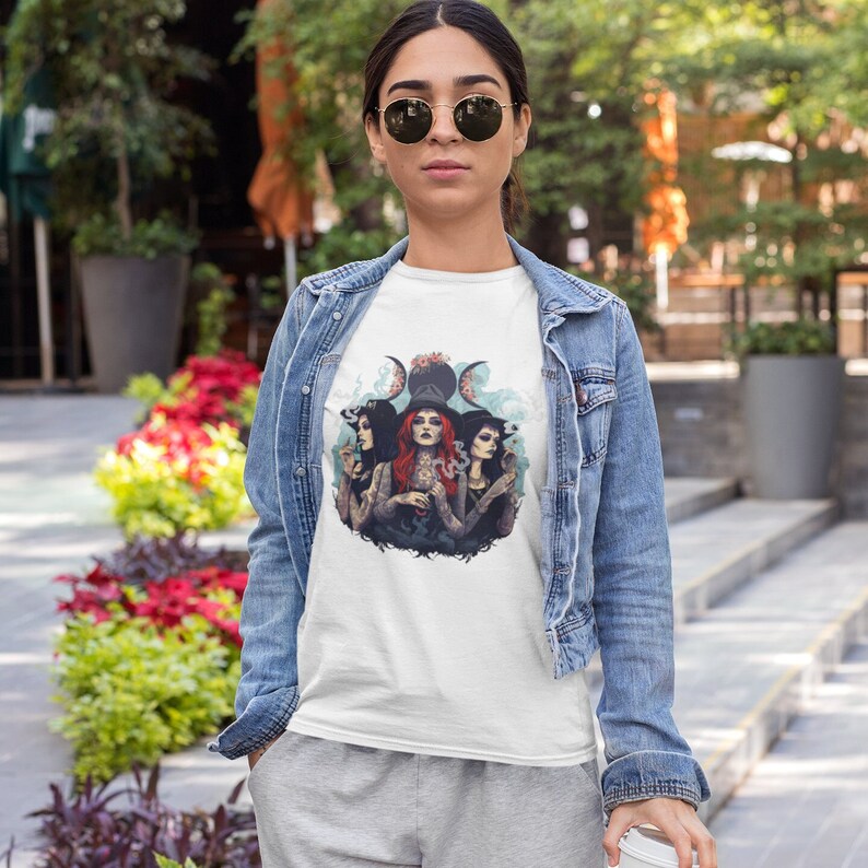 Puede incluir: Camiseta blanca con un gr&aacute;fico de tres mujeres con sombreros de bruja y sosteniendo varitas. Las mujeres est&aacute;n rodeadas por un s&iacute;mbolo de triple luna y humo.