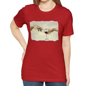 May include: Red t-shirt with a white graphic of two hands reaching towards each other, holding a glass of red wine. The graphic is a parody of the famous "The Creation of Adam" painting.