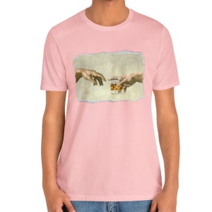 K&ouml;nnte beinhalten: Ein pinkfarbenes T-Shirt mit einem Grafikdruck von zwei H&auml;nden, die sich gegenseitig erreichen und ein Glas Whisky halten. Die Grafik ist eine Parodie auf das ber&uuml;hmte Gem&auml;lde "Die Erschaffung Adams".