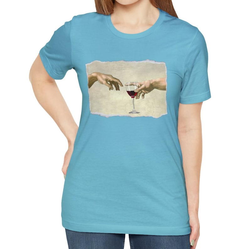 May include: A light blue t-shirt with a graphic of two hands reaching towards each other, holding a glass of red wine. The graphic is inspired by the famous painting "The Creation of Adam".