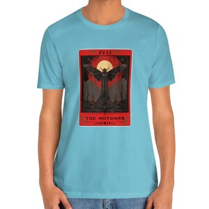 May include: A light blue t-shirt featuring a red and black graphic of a mothman tarot card. The card displays the text "XVIII" and "THE MOTHMAN".