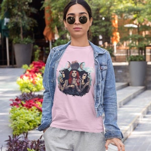 Puede incluir: Camiseta rosa con un gr&aacute;fico de tres brujas con sombreros negros y cabello rojo, de pie frente a un s&iacute;mbolo de triple luna. Las brujas sostienen varios objetos, incluyendo un caldero, una varita y un libro.