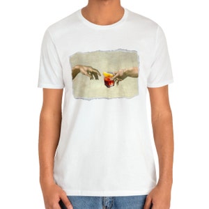 May include: A white t-shirt with a graphic of two hands reaching towards each other, holding a glass of red liquid with a lemon wedge. The graphic is a parody of the famous "Creation of Adam" painting.