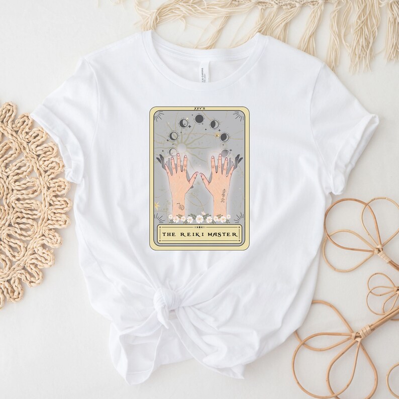 May include: White t-shirt featuring a graphic of a tarot card depicting hands with a moon and stars around them. The card reads "The Reiki Master."