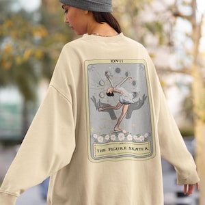 以下が含まれることがあります： フィギュアスケーターのタロットカードのデザインが施されたベージュのスウェットシャツ。カードは、スケーターのイラスト、上部に「XXVII」、下部に「THE FIGURE SKATER」というテキストが入った、明るい灰色の背景です。スウェットシャツはニュートラルカラーです。