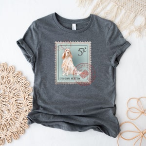 Op de afbeelding: Donkergrijs t-shirt met een postzegelontwerp met een illustratie van een Engelse Setter hond. De postzegel bevat de tekst "5&cent;", "English Setter" en een postdienstembleem. De mouwen van het t-shirt zijn opgerold.