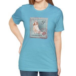 Op de afbeelding: Lichtblauw T-shirt met een postzegelontwerp met een Engelse Setter. De postzegel toont "5&cent;" en "English Setter" met postmarkeringen. Het shirt heeft een effen lichtblauwe kleur.