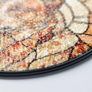 May include: Close-up of a decorative plate with a mosaic design. The plate features a black rim and a surface composed of various shades of orange, red, and cream-colored pieces, creating a textured appearance. The mosaic pieces are outlined with black lines.