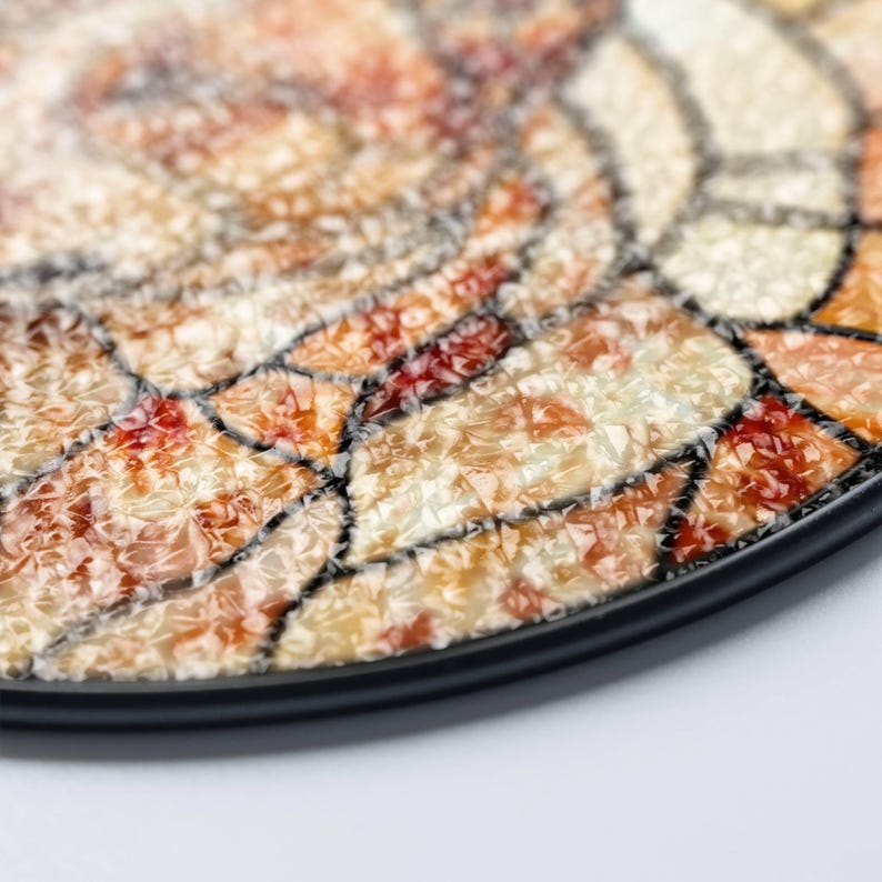 May include: Close-up of a decorative plate with a mosaic design. The plate features a black rim and a surface composed of various shades of orange, red, and cream-colored pieces, creating a textured appearance. The mosaic pieces are outlined with black lines.