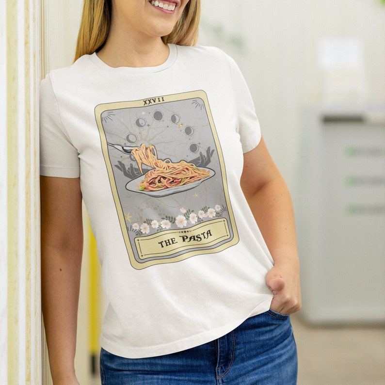 May include: White t-shirt with a graphic of a tarot card design. The card is titled "The Pasta" and features a plate of pasta with a fork and a moon and stars design in the background.
