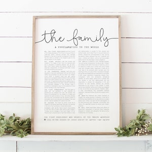 Puede incluir: Una impresión enmarcada con las palabras "the family" en una elegante escritura. Debajo, el texto dice "A PROCLAMATION TO THE WORLD". El marco es de color madera clara y el fondo es blanco. La impresión se muestra en una repisa blanca.