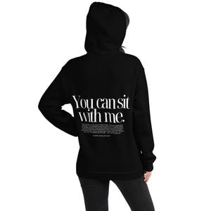 May include: Black hooded sweatshirt with the text "You can sit with me" in white on the back. The sweatshirt also has additional text below the main phrase. The hoodie is a casual garment.