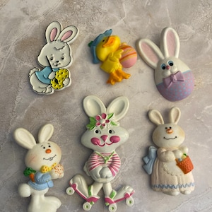 May include: A collection of six Easter-themed decorative figurines. The figurines include white bunnies, a yellow chick, and Easter eggs. The bunnies are in various poses, some holding carrots or wearing outfits. The color palette includes pastels and bright colors.