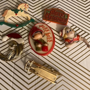 May include: A collection of Christmas-themed pins and brooches. Includes a rocking horse, a Santa hat, a sled, a "Merry Christmas" sign, a figure with "Rum Pa Pum Pum" text, and a figurine of a woman in a red and white outfit.