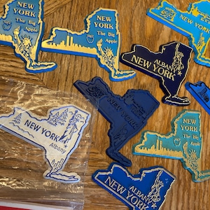 May include: A collection of blue and white New York state souvenir magnets. The magnets are shaped like the state of New York and feature different designs, including the state outline, the New York City skyline, and the Statue of Liberty. Some magnets include the text "New York", "Albany", and "The Big Apple".