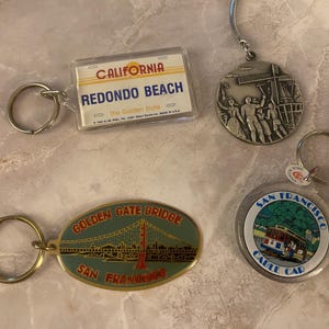 May include: A collection of souvenir keychains featuring iconic California landmarks. Includes a Redondo Beach license plate design, a Golden Gate Bridge oval, a San Francisco cable car, and a windmill scene.