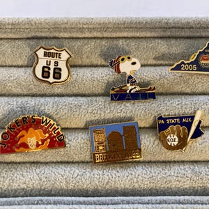 May include: A collection of enamel pins featuring various designs. Included are pins with the Route 66 logo, Snoopy skiing in Vail, a Virginia state pin, a "Joker's Wild" pin, a Breckenridge pin, and a PA State Aux pin.