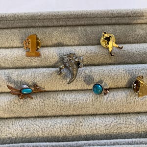 May include: A collection of vintage pins, including a gold number one pin, a silver tornado pin, a gold figure pin, a copper roadrunner pin with a turquoise stone, a blue stone pin, and a gold pyramid pin.