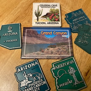 May include: A collection of six green and white Arizona souvenir magnets. The magnets feature different images of the state, including a cactus, a map, and the Grand Canyon. The text on the magnets includes "Arizona", "Phoenix", "Grand Canyon", and "Colossal Cave Mountain Park".