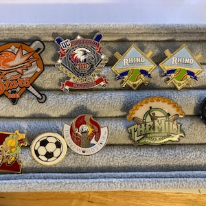 May include: A collection of enamel pins with various designs. Included are pins with baseball bats, a soccer ball, a bicycle, and the text "Let's Go Hawks!". Other pins feature the text "RHINO", "THE MILL", and "OG FREEDOM".