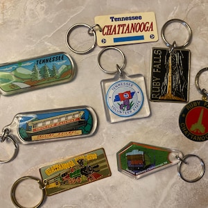 May include: A collection of keychains featuring various designs and the text "Tennessee Chattanooga" and "Ruby Falls". The keychains have metal rings and depict scenes of mountains, trains, and landmarks.