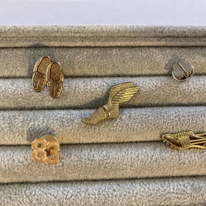 May include: A collection of gold-colored pins, including a pair of running shoes, a winged sandal, a track shoe, and a pin with the number 83. The pins are displayed on a gray, textured surface.