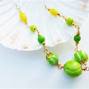May include: A gold necklace with green and yellow glass beads. The beads are in a graduated size, with the largest beads at the bottom of the necklace. The necklace is on a white background.
