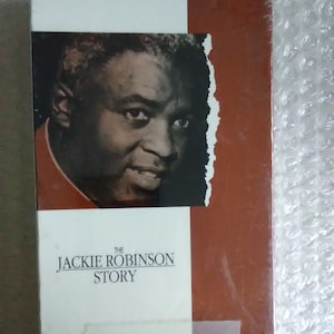 Puede incluir: Una cinta VHS con una cubierta roja y blanca que presenta una foto en blanco y negro de Jackie Robinson. El título dice "The Jackie Robinson Story" y el texto "Starring Jackie Robinson Ruby Dee" está debajo del título.
