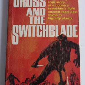 May include: Vintage paperback book cover titled "The Cross and the Switchblade." The cover is predominantly red with white lettering and a black illustration of a person holding a knife. The book is by David Wilkerson.